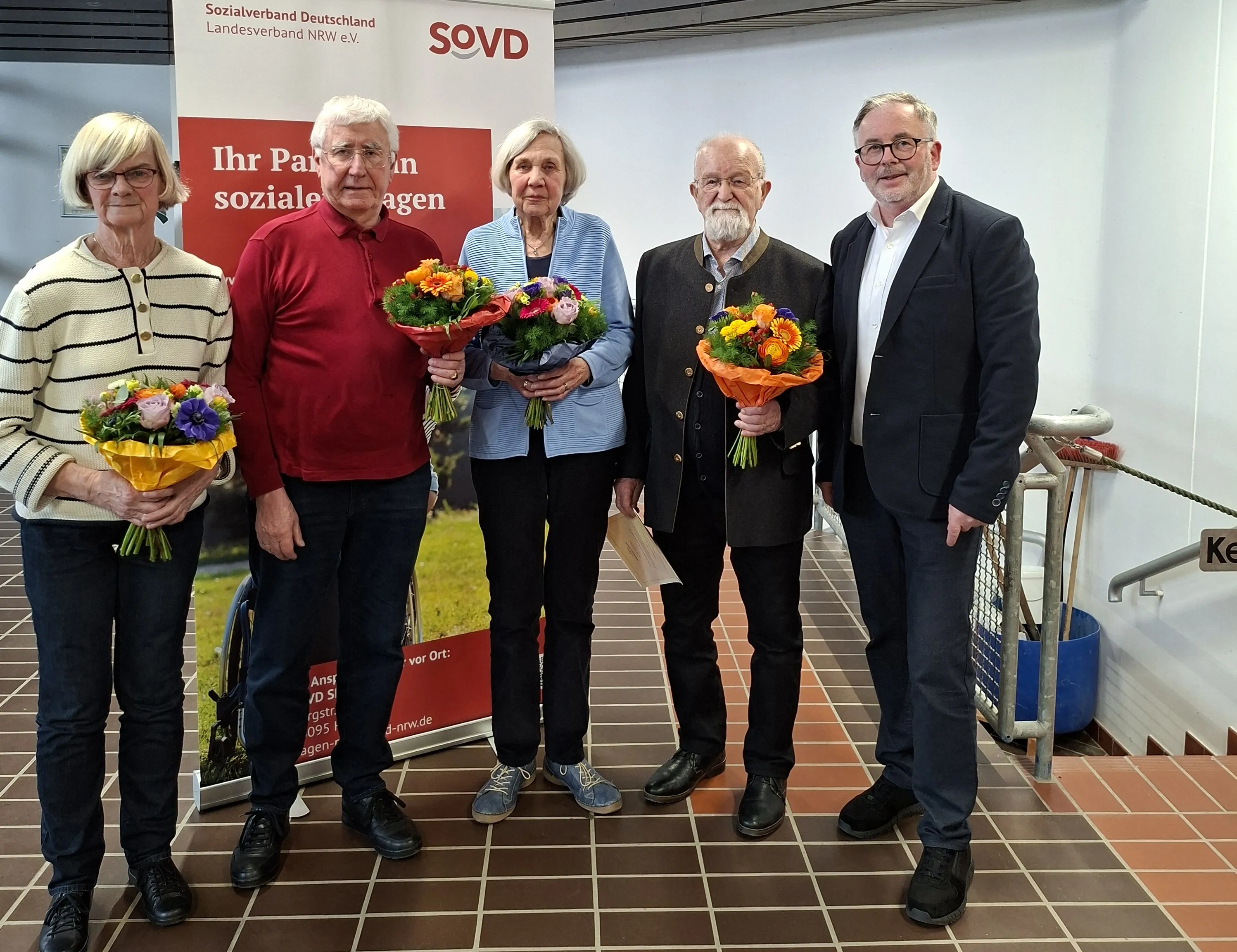 Vier Menschen mittleren und höheren Alters halten Blumensträuße vor einem Banner des Sozialverbands Deutschland. Sie wurden bei einer Mitgliederversammlung geehrt.