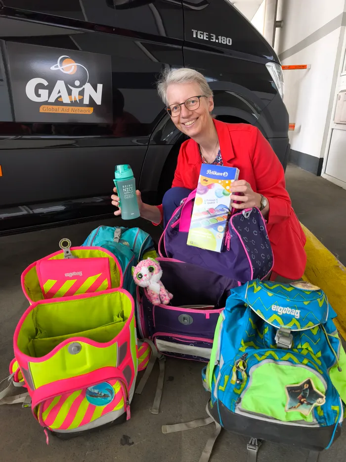 Petra Pientka lächelt vor einem GAIN-Transporter und hält eine Trinkflasche sowie Malutensilien, umgeben von bunten Schulranzen.