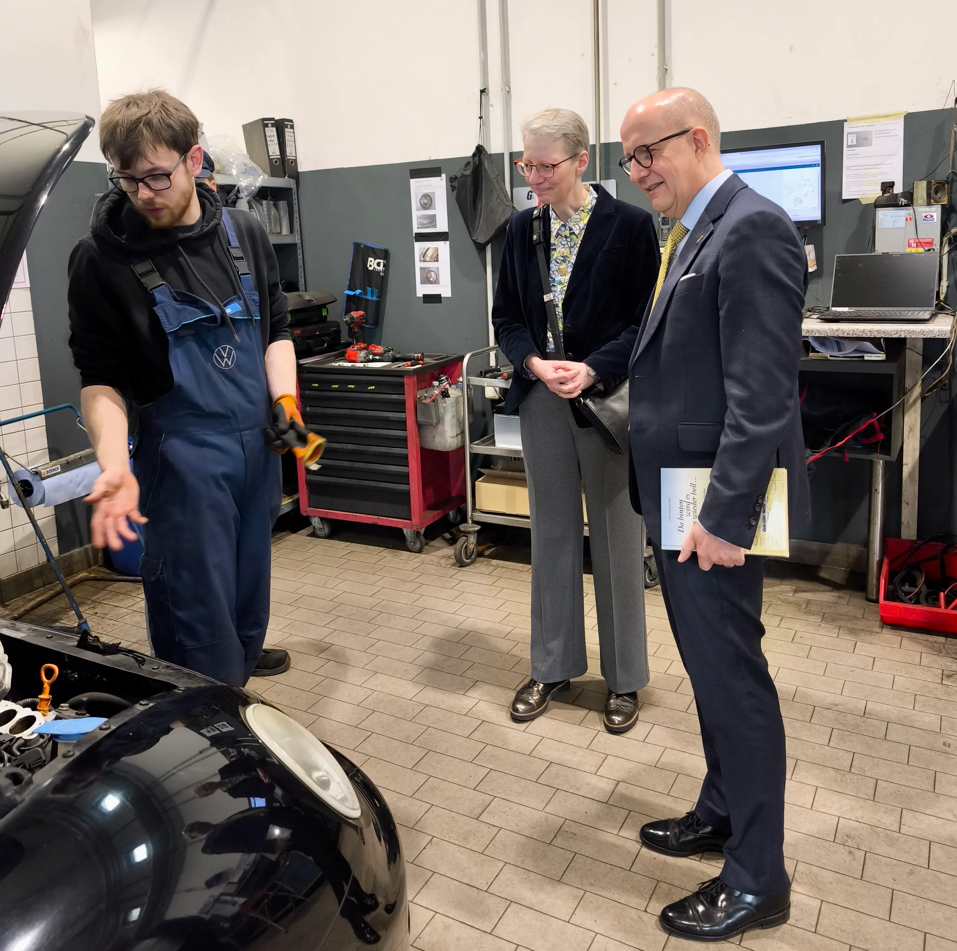 Ein junger Mechaniker mit Schutzbrille zeigt einem Mann im Anzug und einer Frau im Hintergrund die Motorhaube eines Autos.