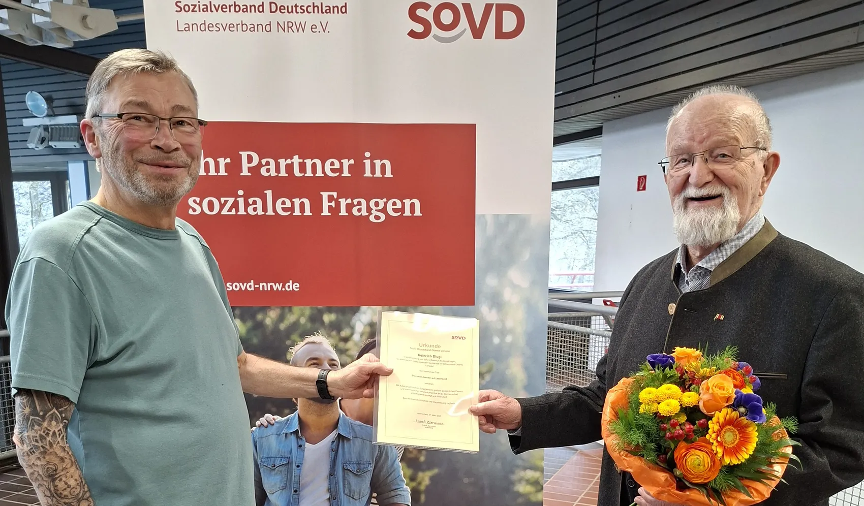 Zwei ältere Männer posieren für ein Foto. Der Mann rechts hält einen Blumenstrauß, während der Mann links eine Urkunde überreicht bekommt, die ein Kind hält.