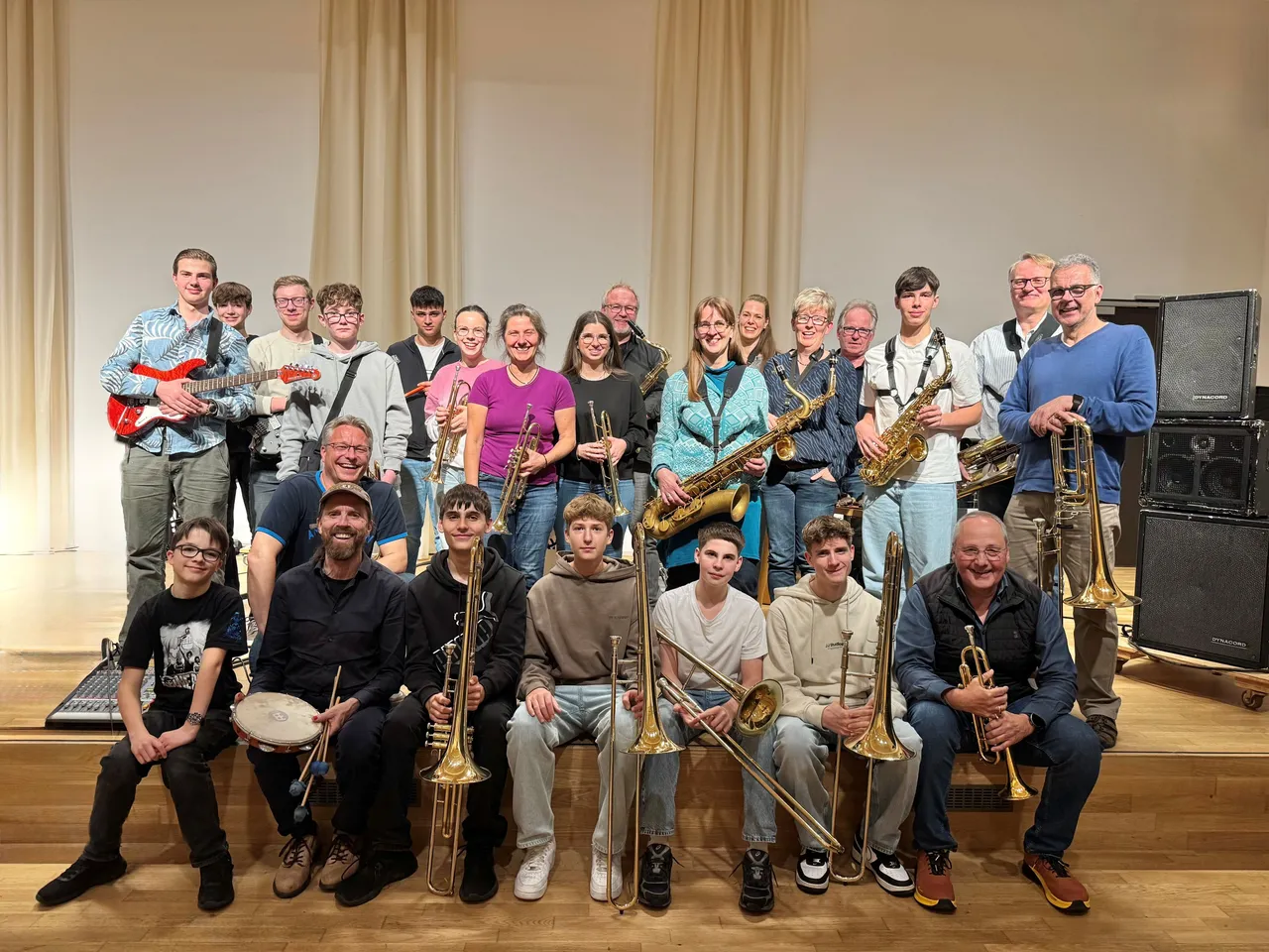 Große Gruppe von Musikern verschiedener Altersgruppen mit Blasinstrumenten und Gitarren posiert auf einer Bühne. Einige Mitglieder der Big Band Footprints SL spielen während ihres Auftritts in der Musikschule.