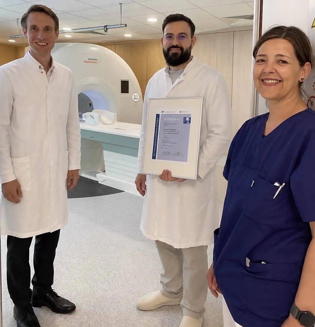 Dr. Thomas Ochtrop, Aladin Thabet und Andrea Rapp stehen vor einem MRT-Gerät und halten ein Zertifikat. Thabet hält das Zertifikat hoch.