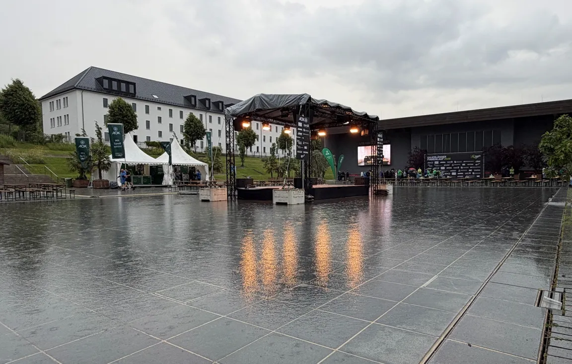 Eine Bühne steht auf einem nassen, gepflasterten Platz vor Gebäuden im Sauerlandpark Hemer. Im Hintergrund sind Zelte und Menschen zu sehen. Die Siegerehrung des Sauerländer AOK-Firmenlaufs wird nachgeholt.