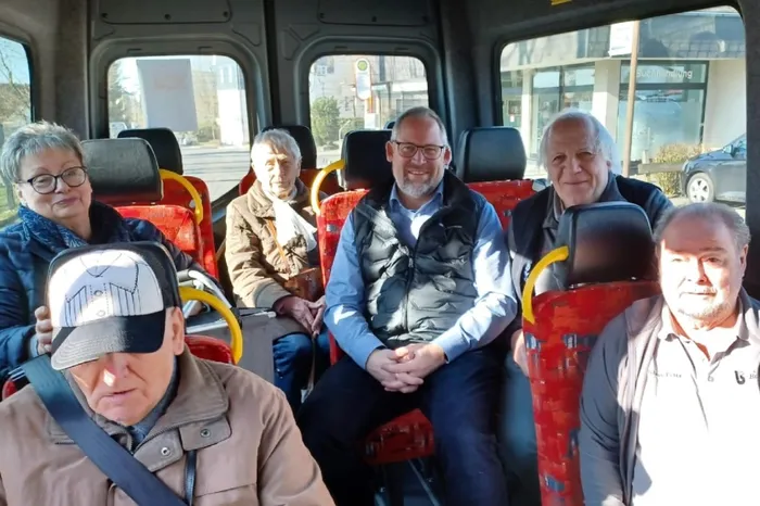 Bürgermeister Armin Kibbert sitzt lächelnd in einem Bürgerbus neben dem Fahrer und weiteren Fahrgästen, darunter der Vereinsvorsitzende. Im Hintergrund ist die Straße durch die Fenster zu sehen.