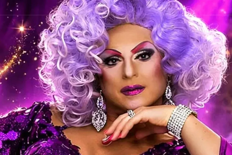 Porträt von Gina de l'Amore mit lilafarbenem Lockenhaar, auffälligem Make-up und funkelndem Schmuck.