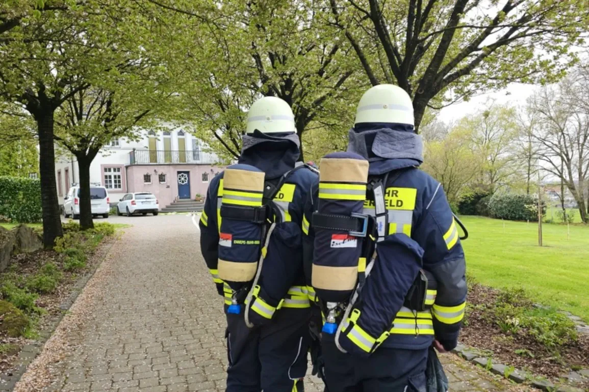 Zwei Feuerwehrleute mit Atemschutzgeräten stehen einander zugewandt, während sie einen Gehweg in einer Wohngegend entlanggehen. Im Hintergrund ein Wohnhaus und parkende Autos.