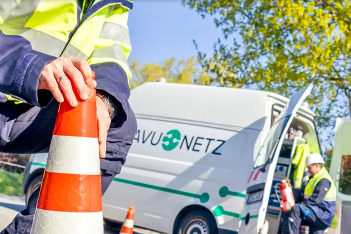Ein Mitarbeiter der AVU Netz in Warnweste hält eine orange-weiße Verkehrskegel vor einem weißen Transporter mit dem AVU Netz Logo.