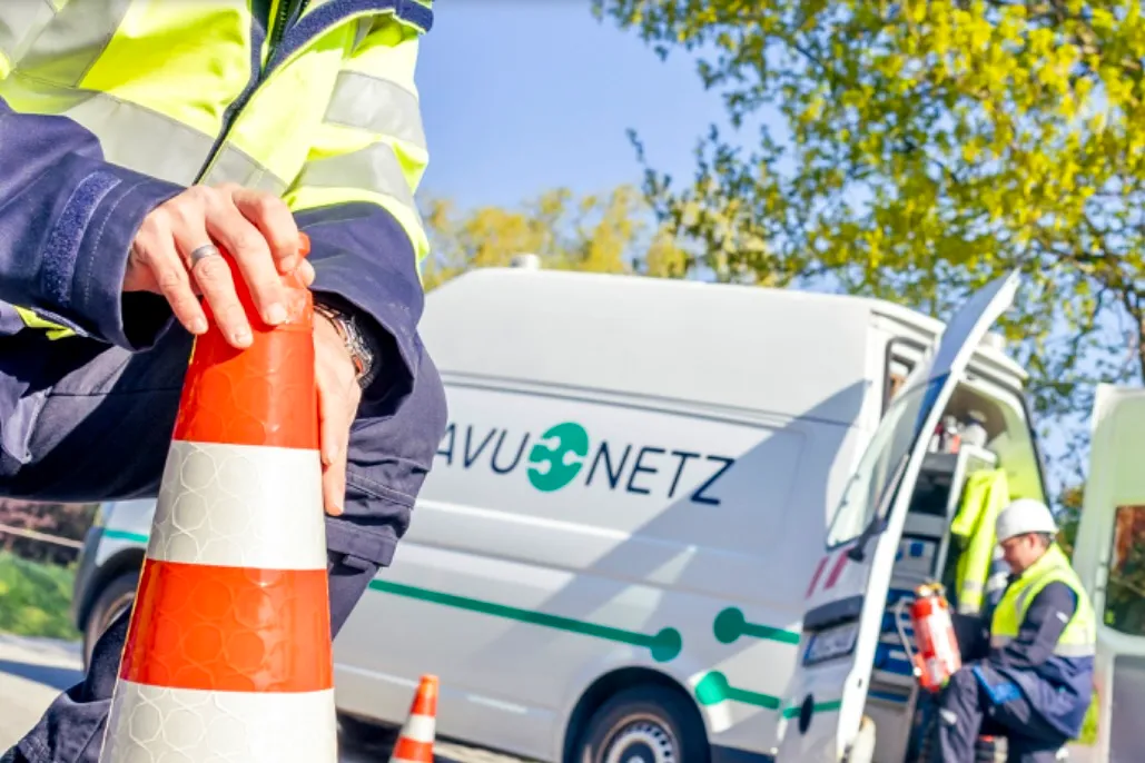 Ein Mitarbeiter der AVU Netz in Warnweste hält eine orange-weiße Verkehrskegel vor einem weißen Transporter mit dem AVU Netz Logo.