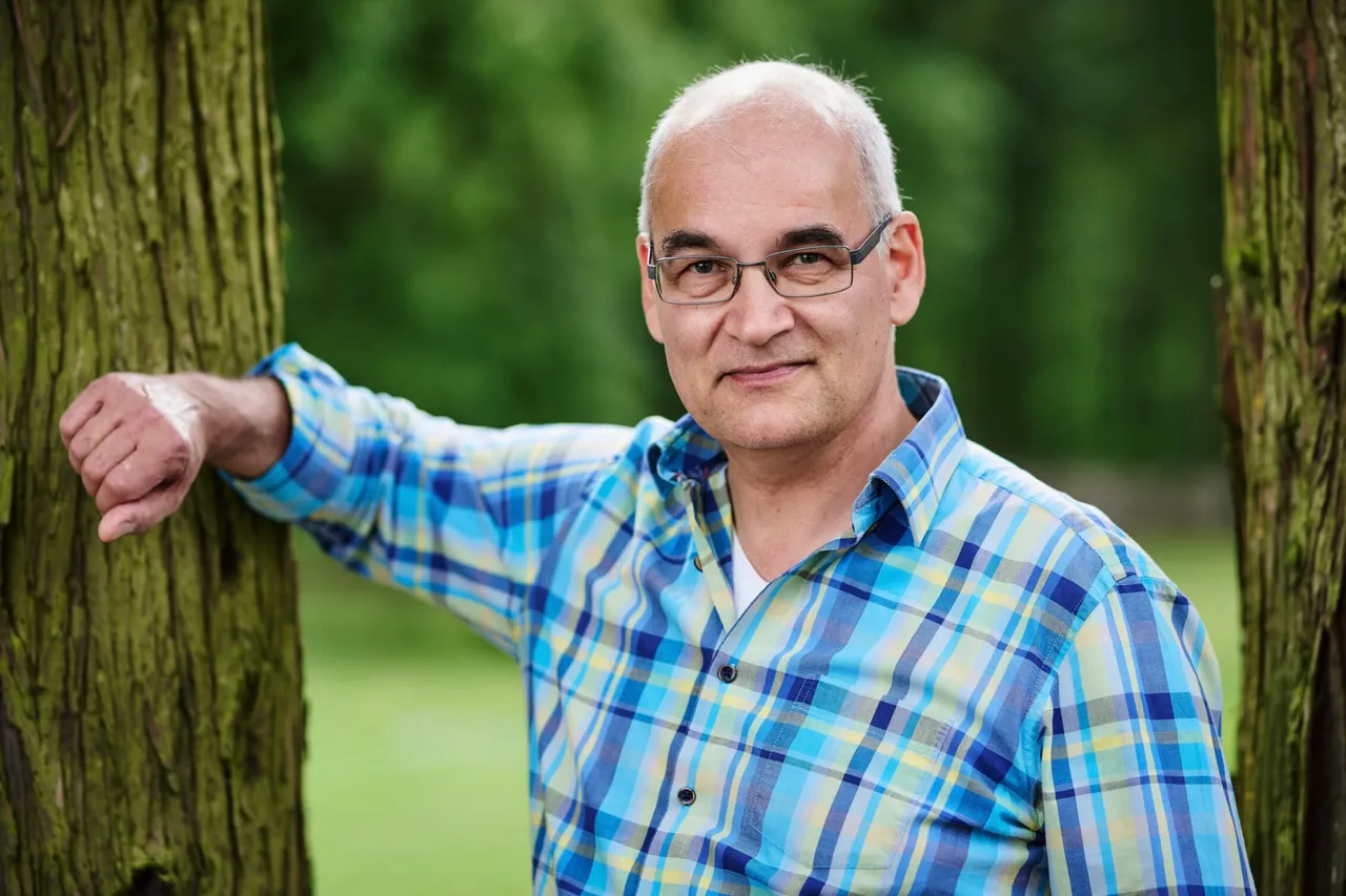 Ein Mann mit Glatze und Brille lehnt lächelnd an einem Baumstamm. Er trägt ein blau-kariertes Hemd und blickt in die Kamera.