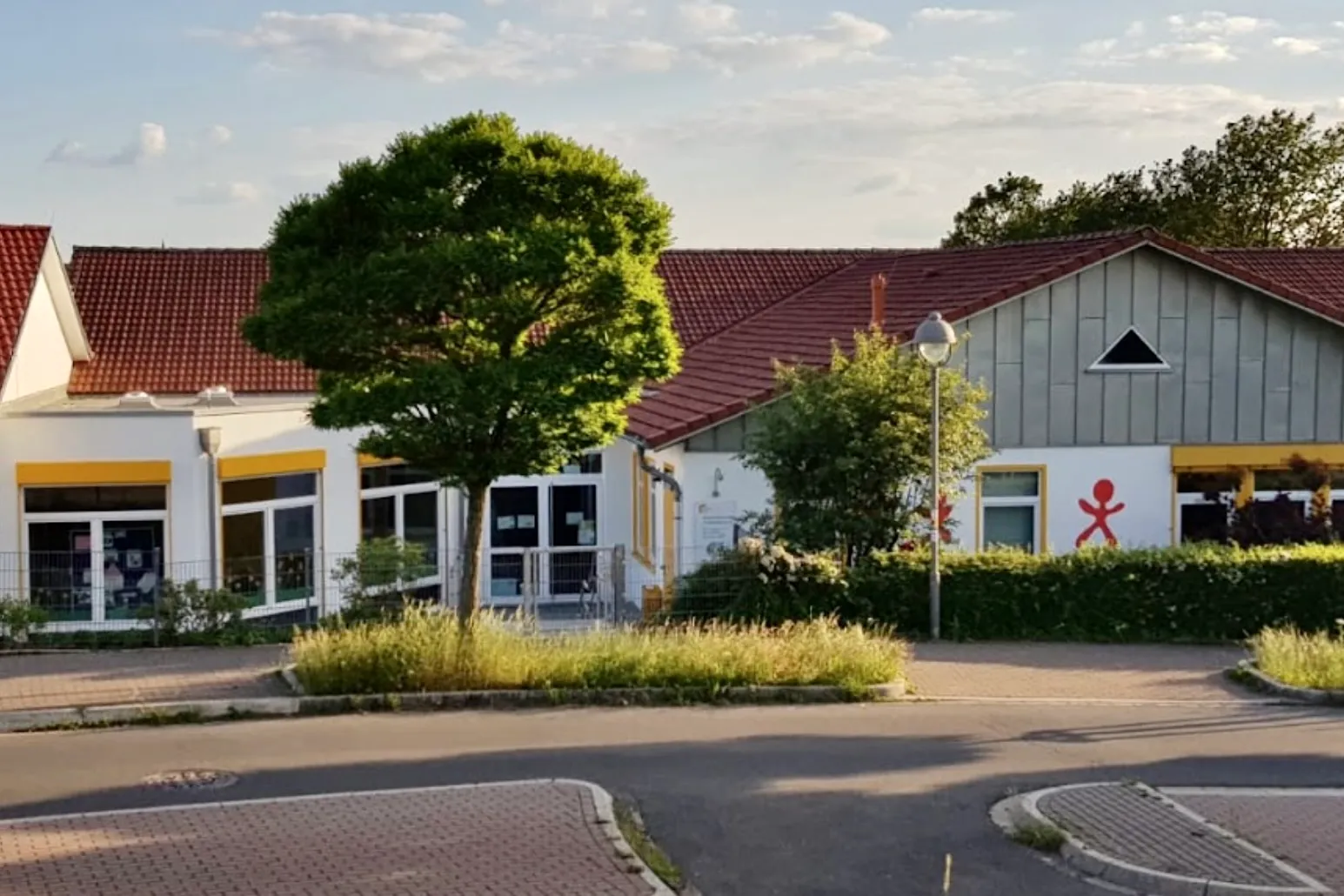 Ein helles, eingeschossiges Gebäude mit einem roten Ziegeldach und gelben Fassadenelementen. Vor dem Gebäude befindet sich ein kleiner, runder Grünstreifen und ein Parkplatz. Ein Kindersymbol ist an der Fassade angebracht.