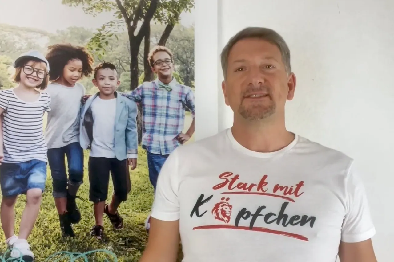 Ein Mann mit kurzgeschnittenem, grau meliertem Haar lächelt in die Kamera. Er trägt ein weißes T-Shirt mit dem Aufdruck „Stark mit Köpfchen“ in roter Schrift. Im Hintergrund ist ein großes Bild von vier Kindern unterschiedlicher ethnischer Herkunft zu sehen, die lachend auf einer Wiese herrennen.