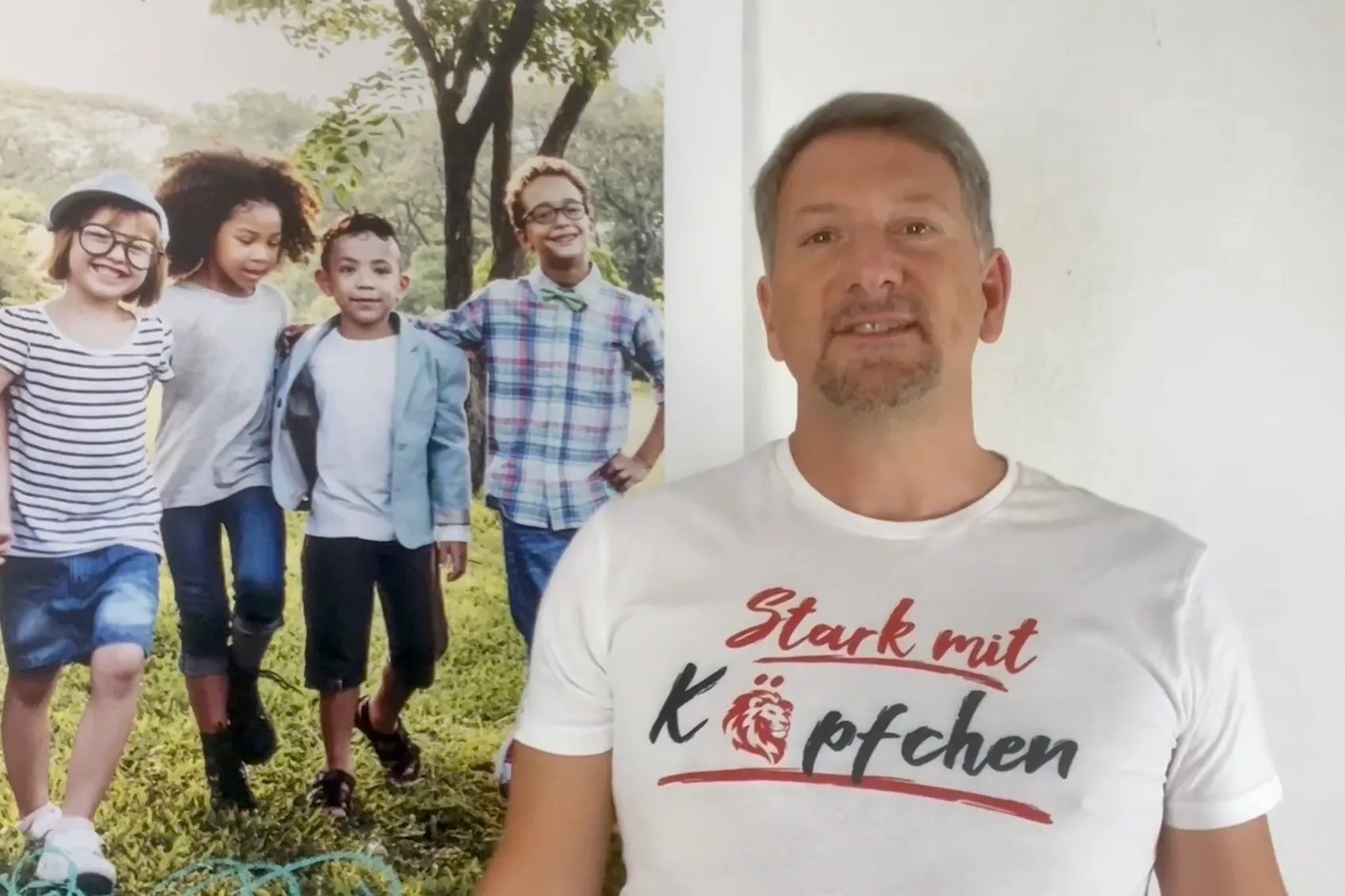 Ein Mann mit kurzgeschnittenem, grau meliertem Haar lächelt in die Kamera. Er trägt ein weißes T-Shirt mit dem Aufdruck „Stark mit Köpfchen“ in roter Schrift. Im Hintergrund ist ein großes Bild von vier Kindern unterschiedlicher ethnischer Herkunft zu sehen, die lachend auf einer Wiese herrennen.