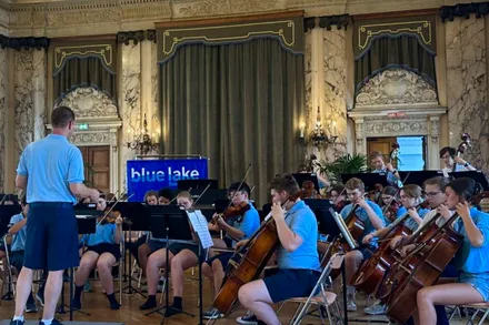 Ein Orchester mit jungen Musikern spielt in einem eleganten Saal. Der Dirigent steht mit dem Rücken zum Betrachter und dirigiert das Orchester, während die Musiker Geigen, Celli und andere Instrumente spielen. Im Hintergrund ist ein Banner mit der Aufschrift „blue lake“ zu sehen.