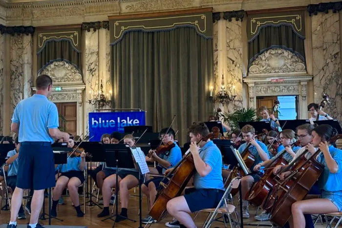 Ein Orchester mit jungen Musikern spielt in einem eleganten Saal. Der Dirigent steht mit dem Rücken zum Betrachter und dirigiert das Orchester, während die Musiker Geigen, Celli und andere Instrumente spielen. Im Hintergrund ist ein Banner mit der Aufschrift „blue lake“ zu sehen.