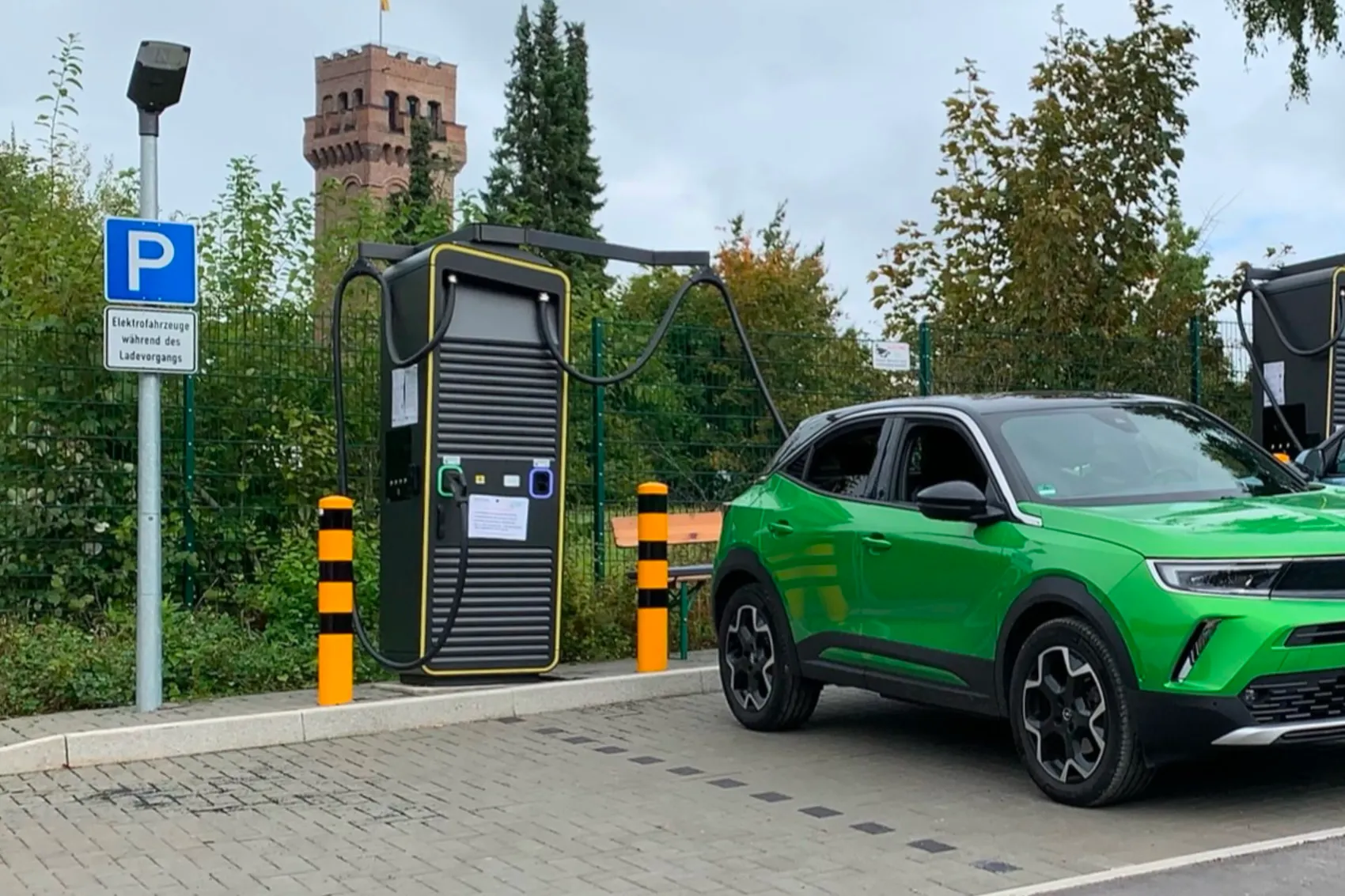 Ein grünes Elektroauto steht an einer schwarzen Ladestation vor Bäumen und einem Backsteinturm.