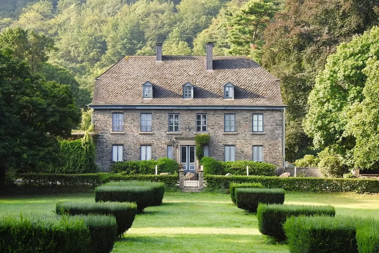 Das zweistöckige Steinhaus Haus Nachrodt mit Gauben vor einem bewaldeten Hintergrund und einem gepflegten Rasengarten mit Formschnitthecken.