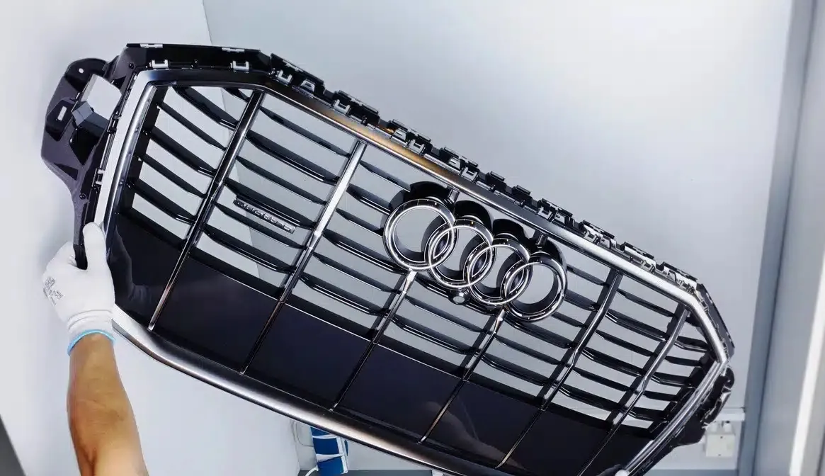 Schwarzer Kühlergrill mit vier silbernen Ringen des Audi-Logos in der Mitte, gehalten von einer Person mit weißen Ärmeln.