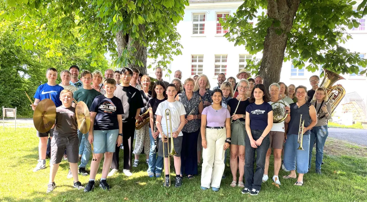 Ein großes Blasorchester mit etwa 40 Mitgliedern unterschiedlichen Alters posiert im Freien vor einem hellen Gebäude unter einem großen Baum. Einige spielen Instrumente wie Posaune, Klarinette und Tuba.