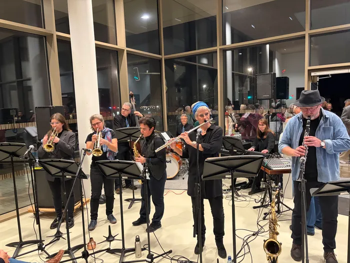 Jazzband Blue Steps spielt in einem hellen Raum. Mehrere Musiker stehen vor Mikrofonen und Notenständern, spielen Trompete, Posaune, Klarinette, Saxophon und Keyboard. Im Hintergrund sind weitere Musiker und ein Schlagzeug zu sehen.