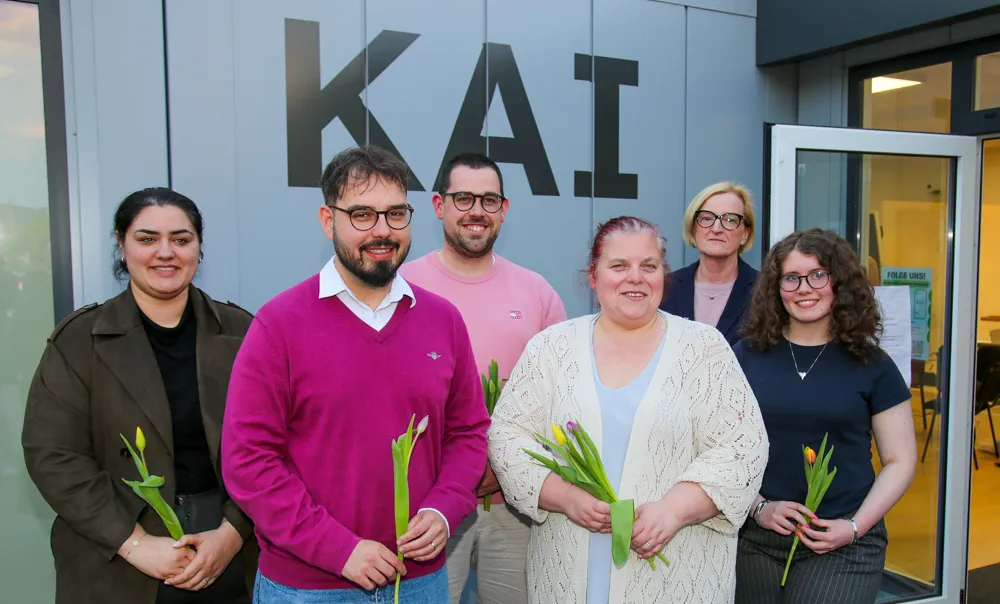 Sechs Personen stehen vor einem Gebäude mit dem Schriftzug „KAI“ und halten gelbe Tulpen. Es sind Jacky Fritz, Jan Schuler, Florian Kraas, Sylvia Olbrich, Petra Hesser und Lilly Mikus, der neue Vorstand der Grünen in Iserlohn.