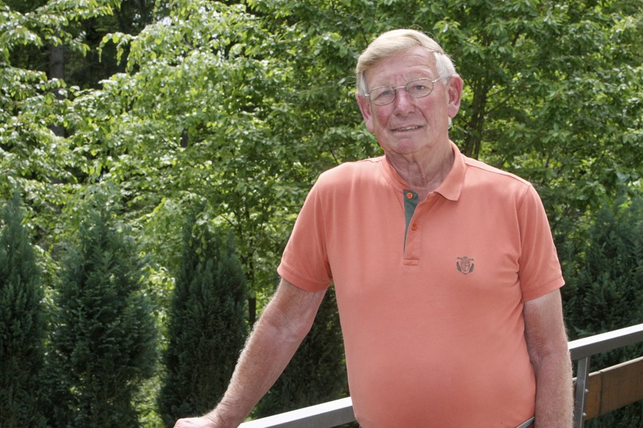 Porträt von Franz-Josef Bomnüter, einem älteren Mann mit Brille und orangefarbenem Poloshirt, vor grünem Hintergrund.