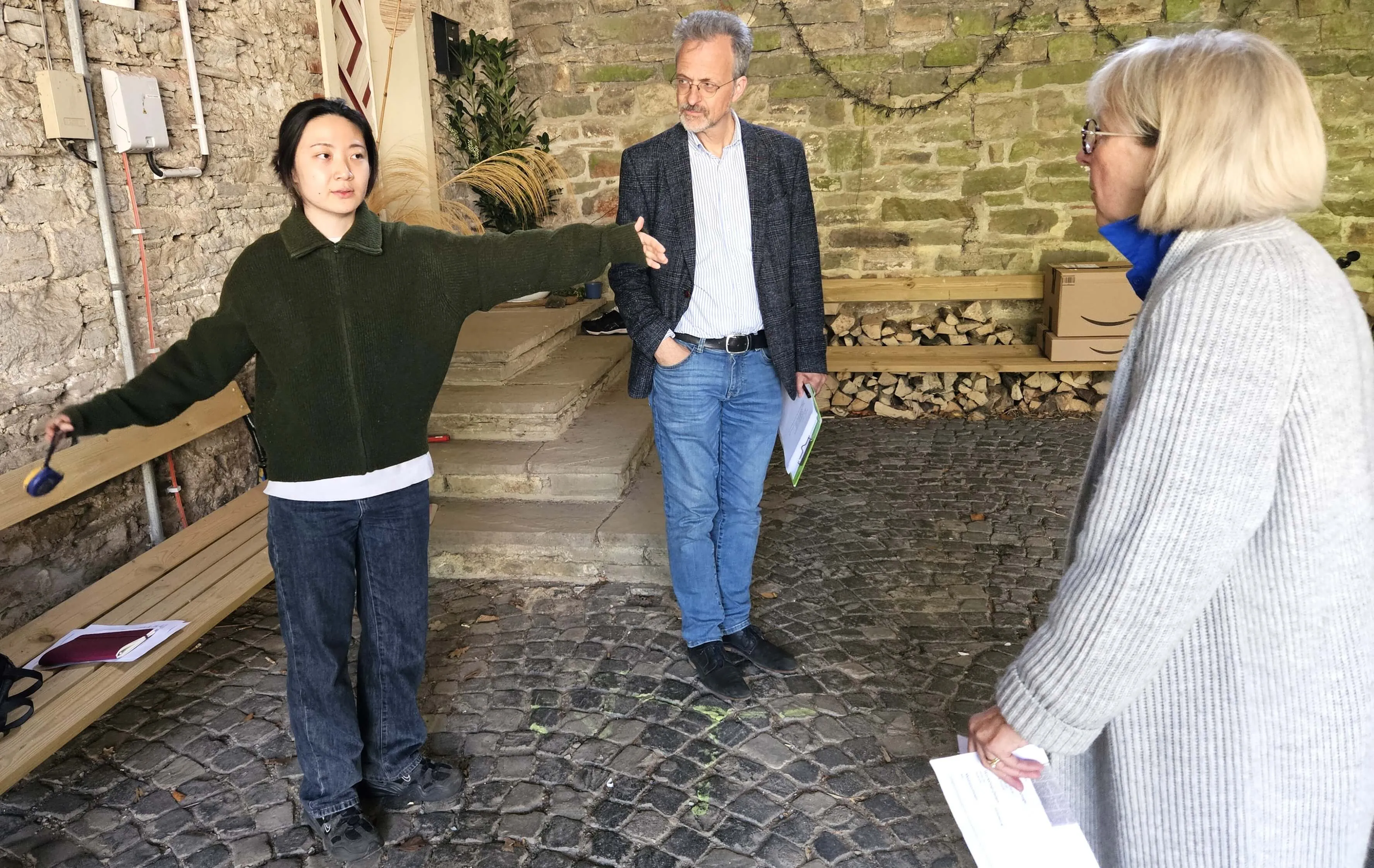 Bon Kim gestikuliert im Gespräch mit Christa und Egbert Cardinal von Widdern vor einer steinernen Mauer. Sie halten Unterlagen in den Händen.