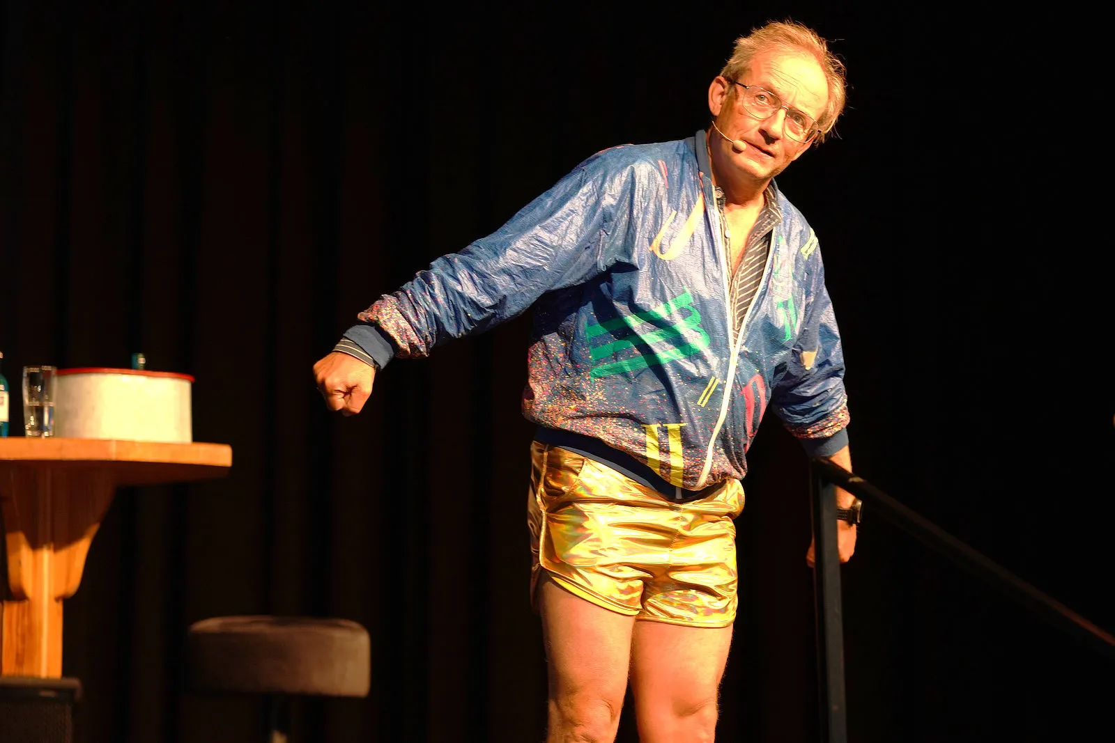 Wigald Boning in bunter Trainingsjacke und goldenen Shorts steht auf einer Bühne und gestikuliert während einer Improvisationsvorstellung.