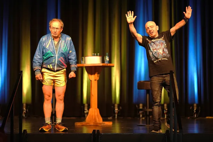 Zwei Comedians auf der Bühne: Wigald Boning in bunter Jacke und goldener Shorts, Bernhard Hoëcker mit hoch erhobenen Armen und schwarzem T-Shirt.
