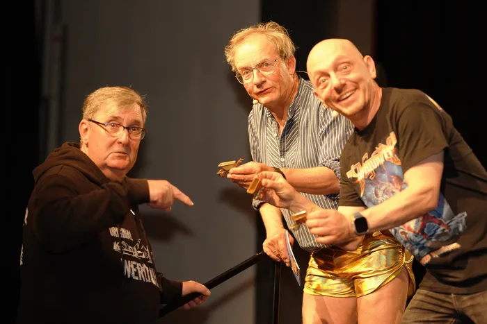 Wigald Boning zeigt mit dem Finger auf Bernhard Hoëcker, der eine goldene Gitarre hält und eine goldene Shorts trägt. Ein dritter Mann lächelt in die Kamera.