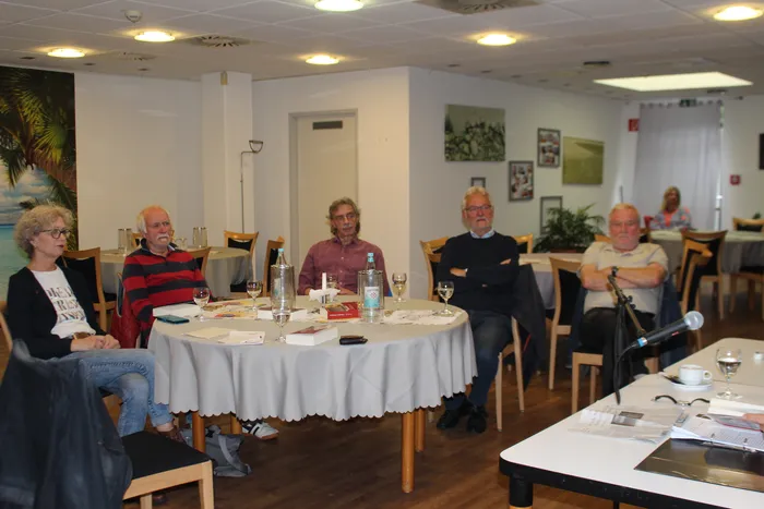 Fünf Personen sitzen an einem runden Tisch mit weißer Tischdecke in einem hellen Raum. Sie hören einem Mann zu, der am Tisch sitzt und spricht. Auf dem Tisch stehen Gläser und Wasserflaschen.