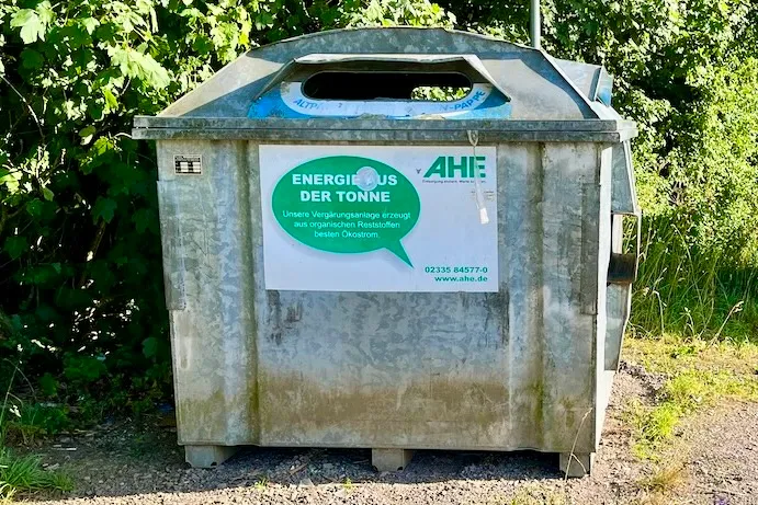 Grauer Altpapiercontainer mit Aufkleber der AHE, der die Energiegewinnung aus Altpapier bewirbt.