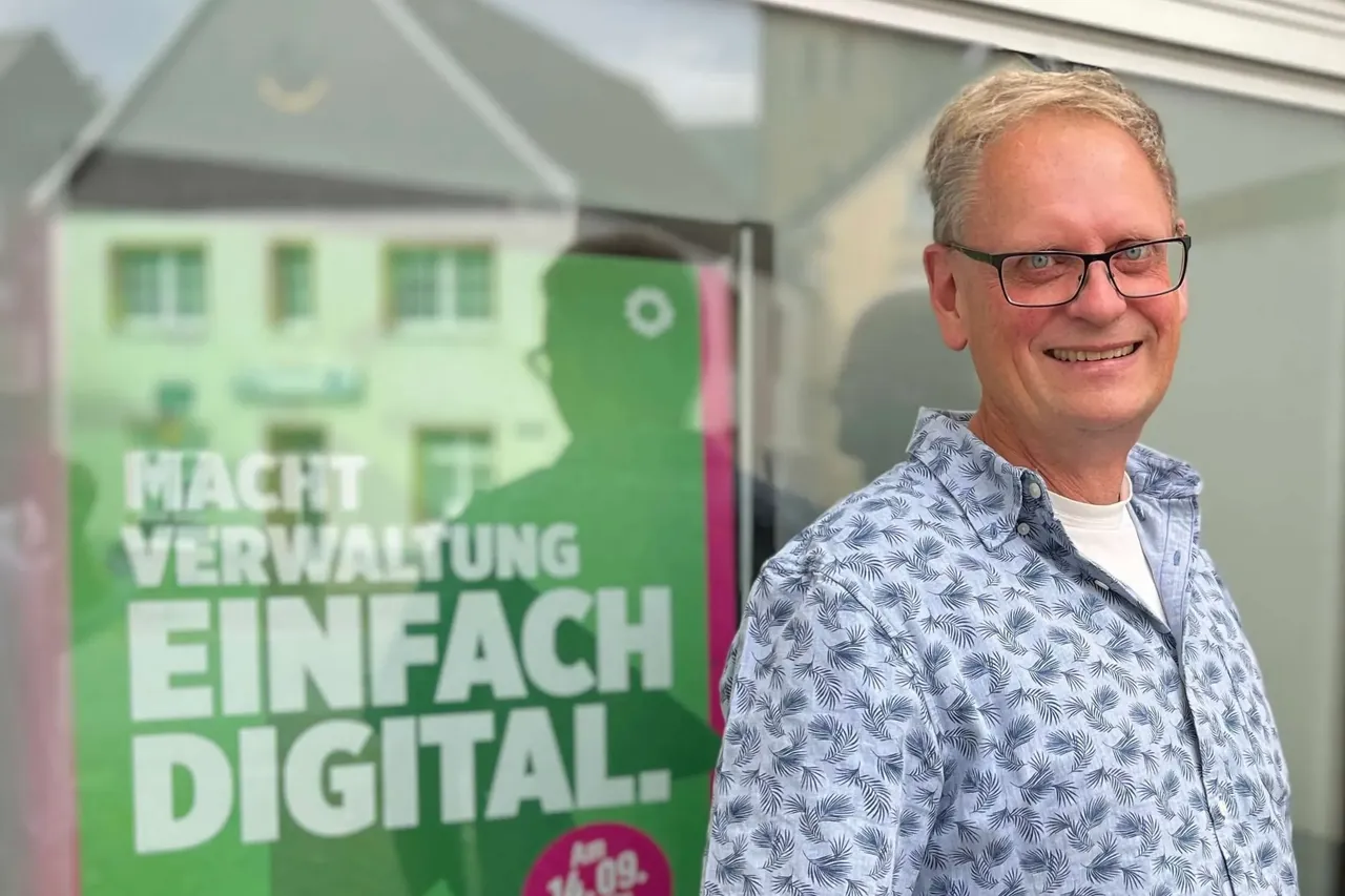 Ein lächelnder Mann mit Brille und blauem Hemd steht vor einem Plakat mit der Aufschrift „Macht Verwaltung einfach digital.“ und einem stilisierten Haus.
