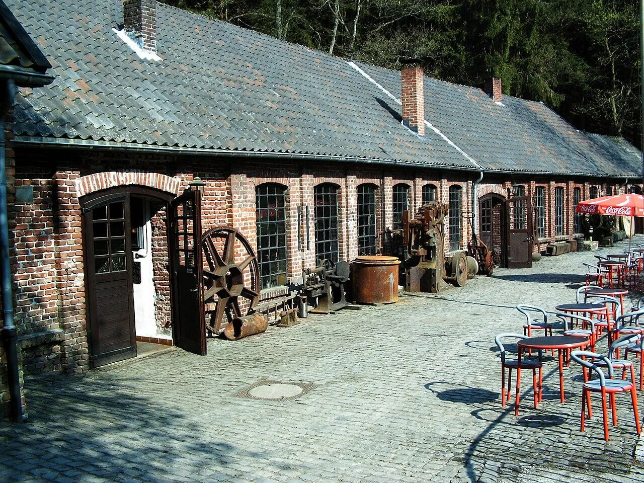 Ein Backsteingebäude mit Ziegeldach, vor dem auf einem gepflasterten Hof alte Industriemaschinen und rote Café-Möbel stehen.