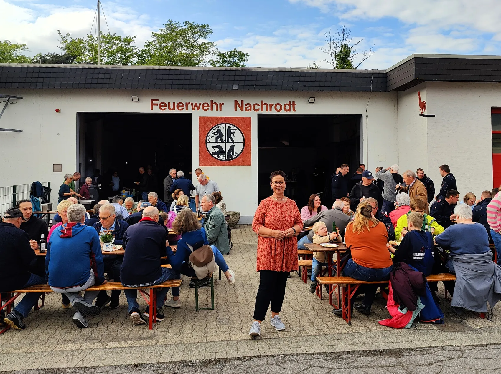 Bürgermeisterin Birgit Tupat steht vor dem Feuerwehrhaus in Nachrodt-Wiblingwerde, umgeben von vielen Menschen bei einem Grillfest.