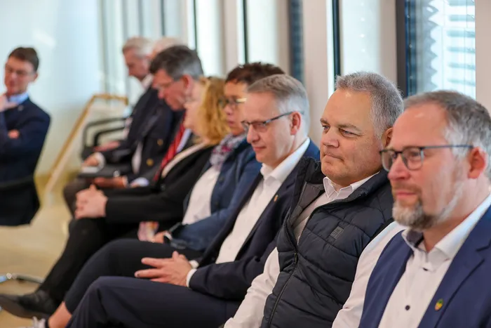 Reihe von Männern und Frauen in formeller Kleidung sitzen auf Stühlen und verfolgen eine Diskussion. Im Vordergrund ein Mann mit grau meliertem Haar und dunkler Jacke.