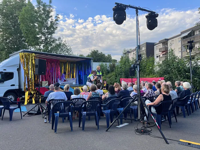 Ein Publikum sitzt auf blauen Stühlen vor einem umgebauten Lastwagen, der als Bühne dient. Auf der Bühne stehen drei Personen, während zwei weitere Personen im Hintergrund zu sehen sind.