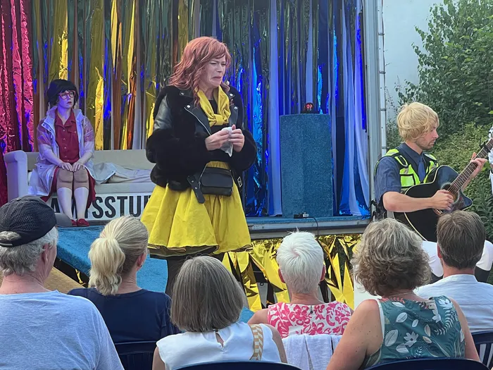 Eine Frau mit roten Haaren steht auf einer Bühne und spricht in die Menge. Im Hintergrund spielt ein Mann Gitarre, links sitzt eine weitere Person.
