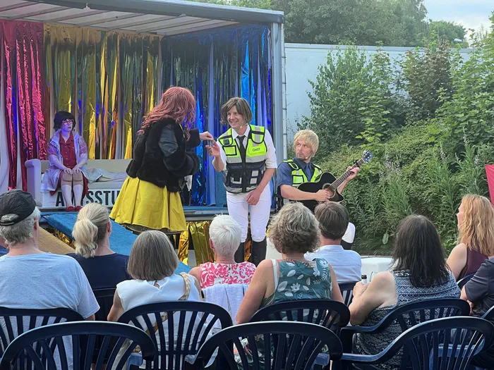 Eine Frau mit roten Haaren spricht in ein Mikrofon, während eine andere in einer gelben und schwarzen Kleidung daneben steht. Im Hintergrund spielen zwei Musiker auf einer Bühne vor einem Publikum.