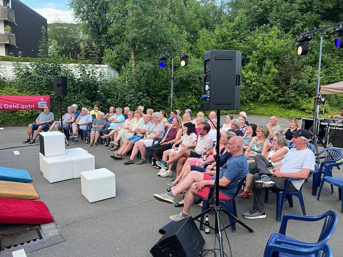 Zuschauer sitzen auf Stühlen und provisorischen Sitzgelegenheiten vor einer Bühne mit Lautsprechern und beobachten eine Theateraufführung im Freien. Im Hintergrund sind Bäume und ein Gebäude zu sehen.