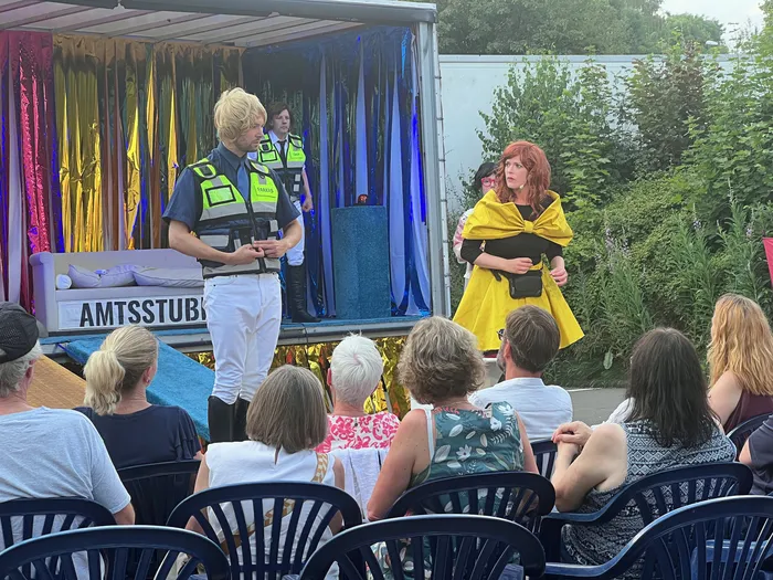 Zwei Schauspieler in Kostümen stehen auf einer Bühne vor einem Publikum. Der Mann trägt eine Uniform und die Frau ein gelbes Kleid. Im Hintergrund steht ein Schild mit der Aufschrift 