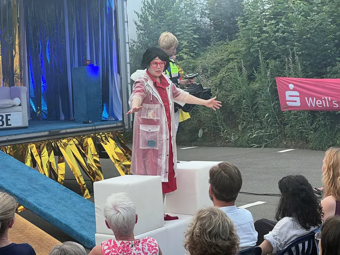 Eine Frau mit Brille, rotem Oberteil und pinkfarbenem Regenmantel steht auf einer Bühne und gestikuliert. Im Hintergrund sitzen Zuschauer und ein Bühnenbild mit dem Schriftzug 