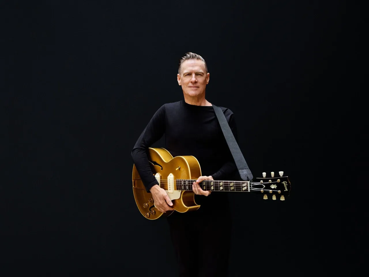 Bryan Adams in schwarzer Kleidung hält eine goldene E-Gitarre vor einem schwarzen Hintergrund.
