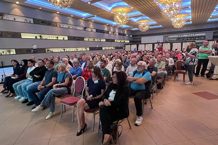 Ein großes Publikum sitzt in Reihen in einem hell erleuchteten Saal mit Kronleuchtern.