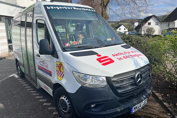 Weißer Bürgerbus mit rotem Streifen und dem Logo des Bürgerbusvereins, geparkt vor einer Hecke und einem Wohnhaus. Der Bus ist mit 'bürgerbus Herscheid e.V.' beschriftet und trägt das Slogan 'Aktiv für alle. In der Region.'.