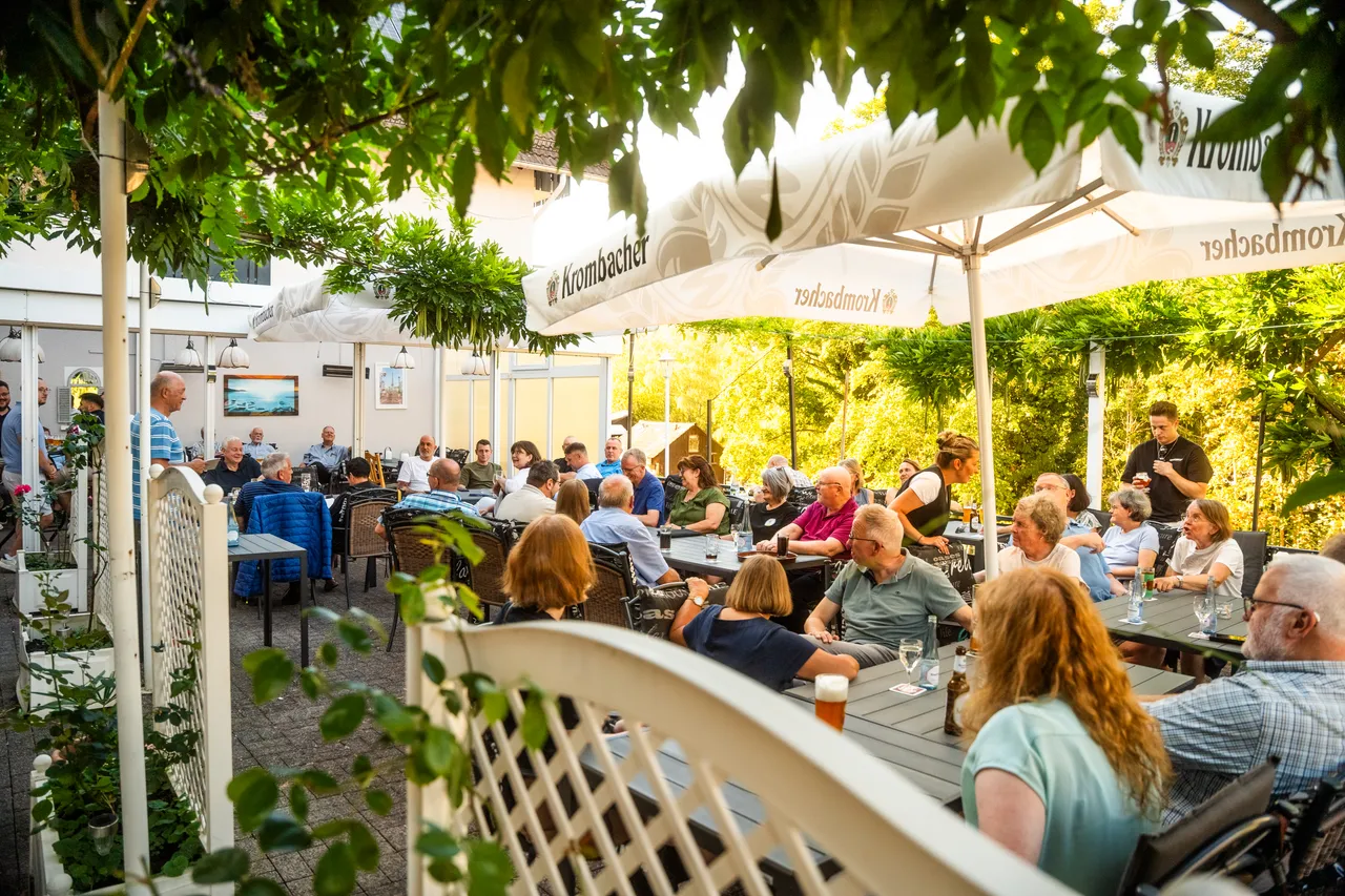 Menschen sitzen an Tischen in einem Biergarten unter großen weißen Sonnenschirmen und unterhalten sich.