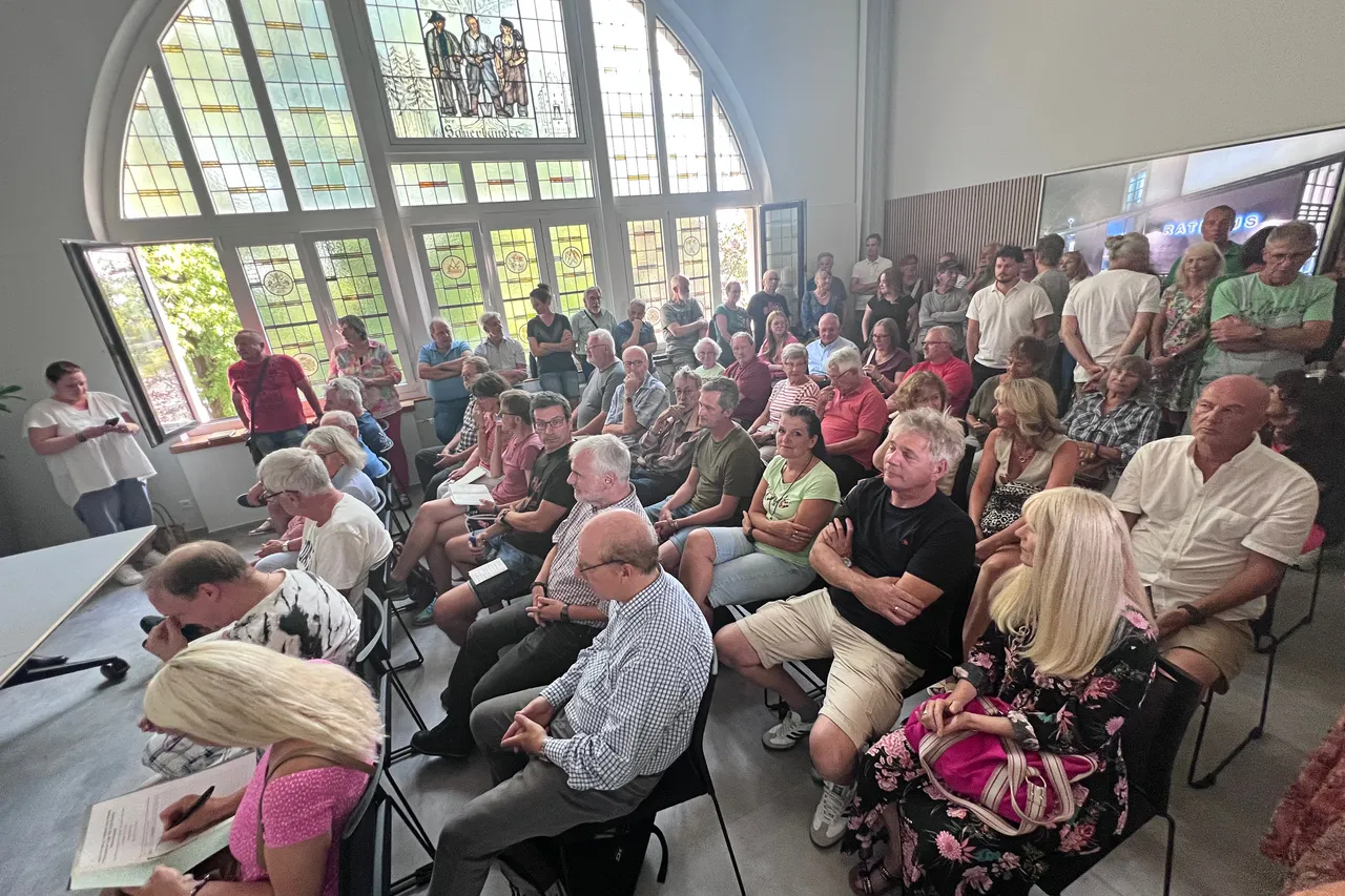 Eine große Gruppe von Menschen sitzt in einem hellen Saal mit großen Bogenfenstern bei einer Bürgerversammlung.