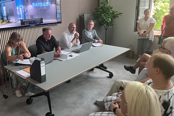 Eine Gruppe von Menschen sitzt an einem Besprechungstisch in einem Raum mit einem Bildschirm, auf dem „RATHAUS“ steht.