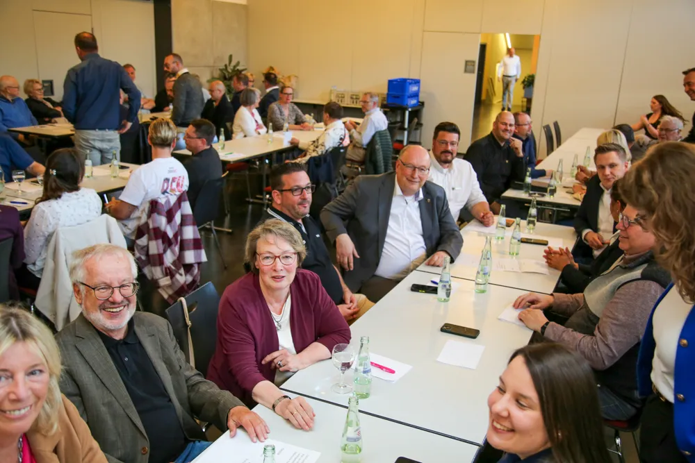 Versammlung von Menschen in einem hellen Raum, viele sitzen an Tischen und schauen in unterschiedliche Richtungen. Im Vordergrund ein lächelnder älterer Mann und eine Frau mit rotem Oberteil.