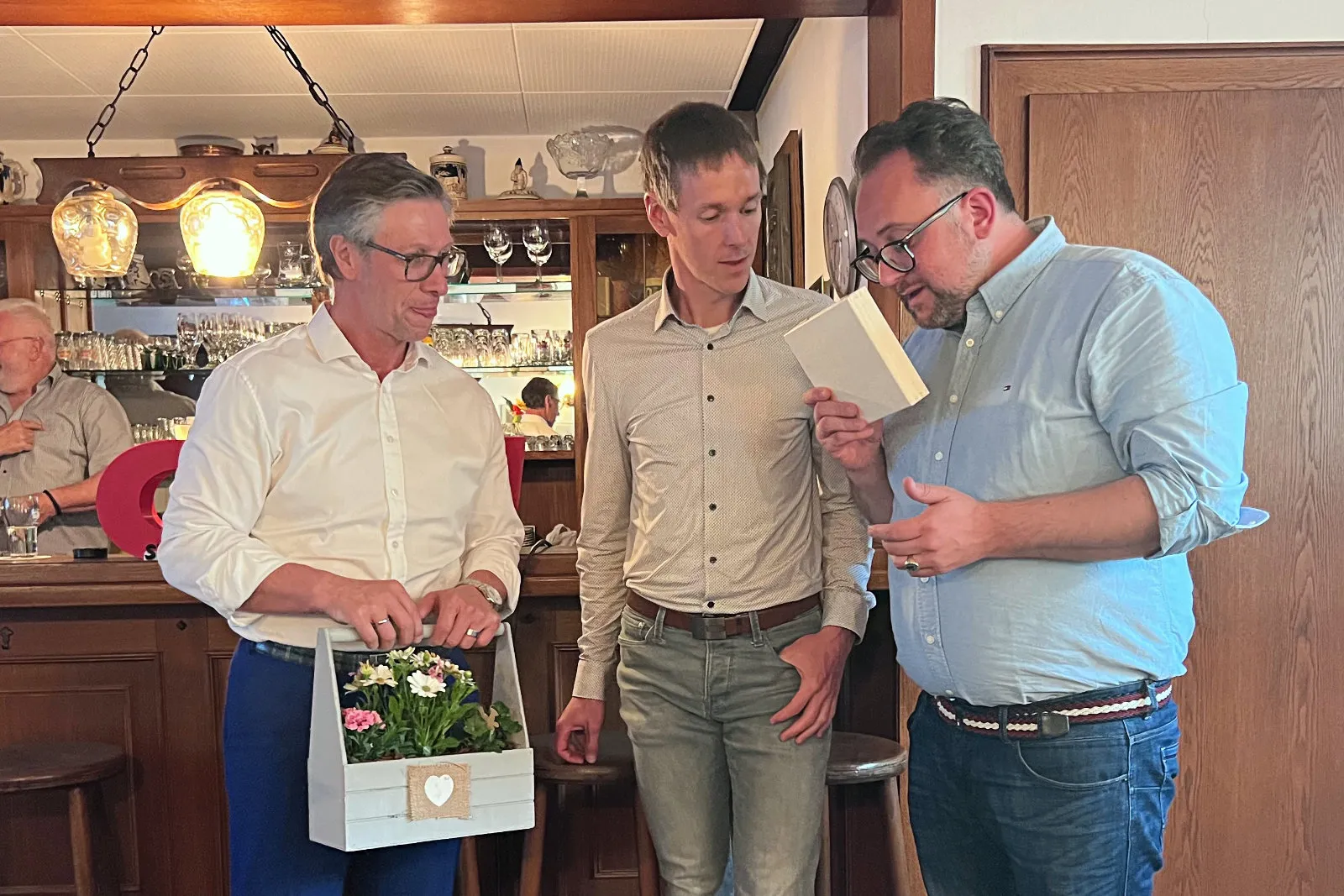 Drei Männer betrachten eine Karte in einer Kneipe. Links hält einer eine Blumenschachtel, im Hintergrund sind Gläser und dekorative Gegenstände zu sehen.