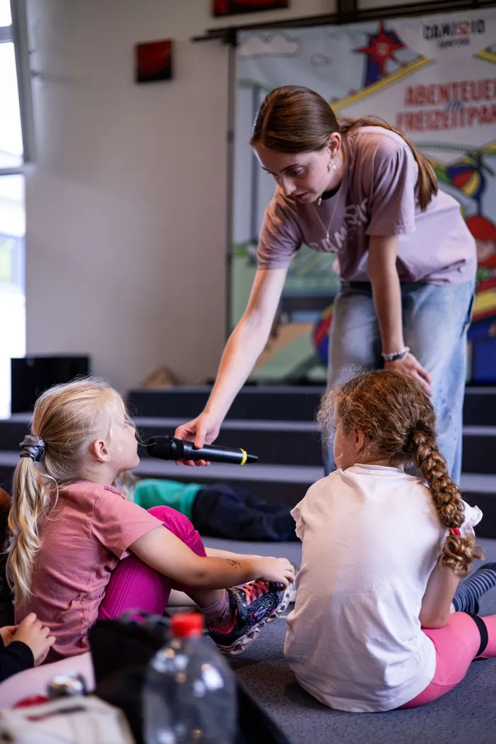Eine junge Frau beugt sich zu sitzenden Kindern hinunter und reicht einem Mädchen ein Mikrofon.